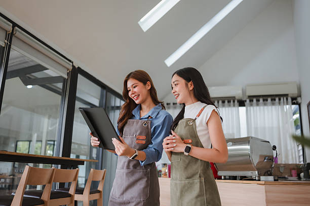 istock Two smiling baristas in aprons discussing menu options and business plans inside a modern coffee shop, using a tablet 2243410790