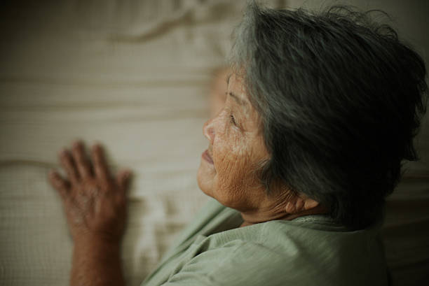 Asian elderly woman lying in bed looking out the window thoughtfully.