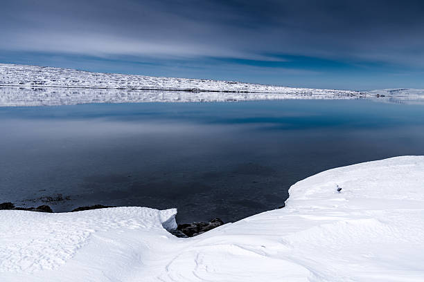 Winter scene with a snow-covered lake and cloudy sky. Ice and snow blanket the landscape. Calm water reflects the surrounding environment.