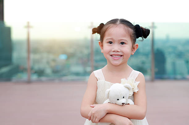 A cute little girl smiling happily while hugging her white teddy bear on a rooftop overlooking the city at sunset.