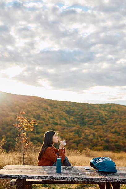 Woman sitting at picnic table, enjoying a warm drink with steam rising, surrounded by autumn nature and golden light