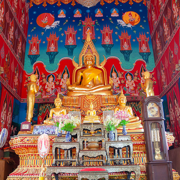 Golden thai buddha in small temple in Bangko,, name un known. Located in Phra Nakhon district. Below buddha statue are some smaller buddha statues and a photo of King Bhumipol with global royals at meeting in Throne Hall in year 2006. In background is  colorful thai art mural. Two golden buddha statues are standing at both sides. There is  also a clock