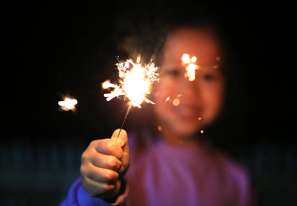 Joyful Asian little girl smiling brightly and holding sparkling, burning firework (sparkler) in the dark, capturing the pure excitement and magic of a festive night celebration.