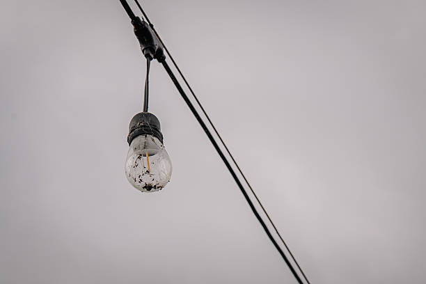 istock Hanging light bulb with visible imperfections and wires against a cloudy sky, showcasing industrial design and atmospheric lighting effects 2243442987
