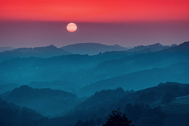 Sun descending over rolling hills, creating a tranquil atmosphere with a red sky and atmospheric haze