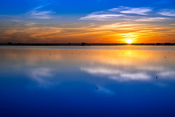 Sunset casts an orange glow across the still waters of Isla Mayor, reflecting wispy clouds and silent harvested rice fields in Andalusia.