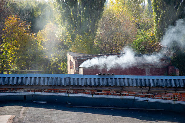 Smoke billows from a red-brick chimney, contrasting with vibrant fall foliage under clear blue skies in a peaceful area.