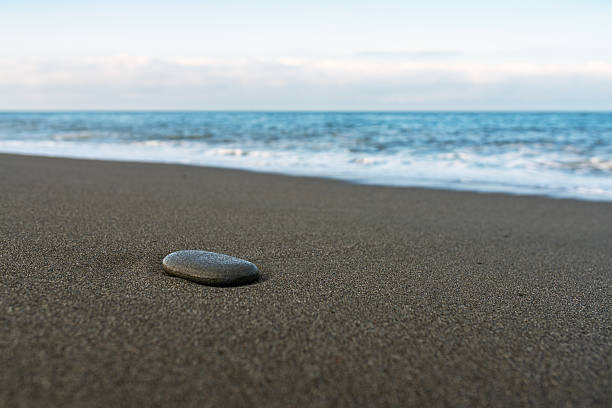 A tranquil beach composition featuring a lone pebble on dark sand, gentle waves, and a distant horizon, evoking calm, solitude, and natural simplicity.