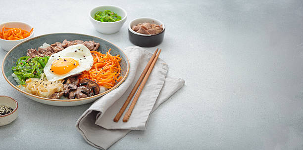 Traditional korean bibimbap with fried egg, beef and vegetables and other toppings on light background with chopsticks, space for text