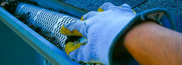 Panorama view handyman in gloves inspecting and clearing debris from white metal gutter guard on dark-shingled roof in Dallas suburb, preparing for seasonal rainfall, DYI home maintenance