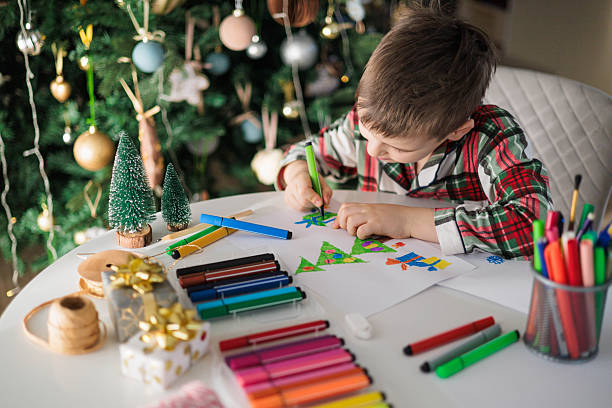 istock Boy Drawing Christmas Tree with Markers at Table near Decorated Holiday Tree 2243481724
