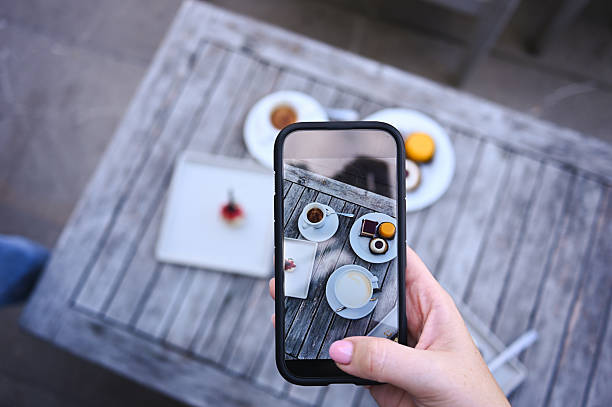 Hand holding a smartphone taking a flat lay image of food and coffee on a wooden table