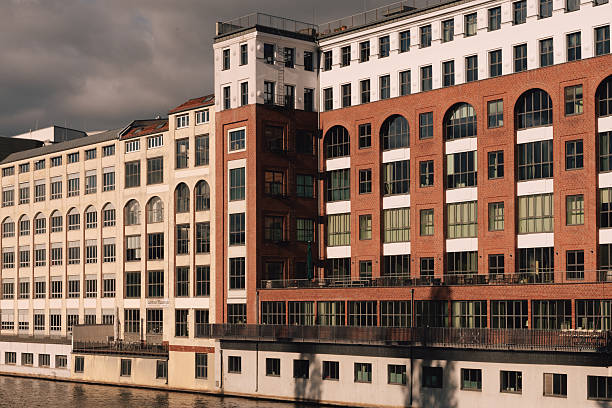 Modern riverside architecture in Berlin, Germany, combining red and white brick facades with large glass windows. The building stands along the Spree River under warm sunlight, emphasizing geometry and reflection.