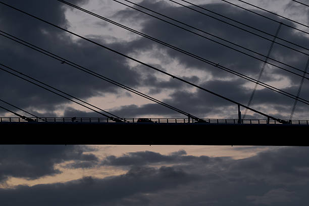 A car crosses a modern cable-stayed bridge silhouetted against a moody evening sky. The intersecting lines of the suspension cables and the dark clouds create a powerful graphic composition that emphasizes geometry, movement, and contrast.
