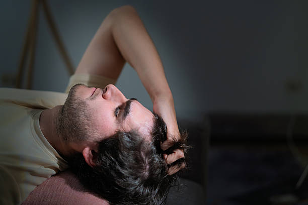 Young man lying on a couch in a dim room, appearing sad and struggling with mental health, pulling his hair in despair