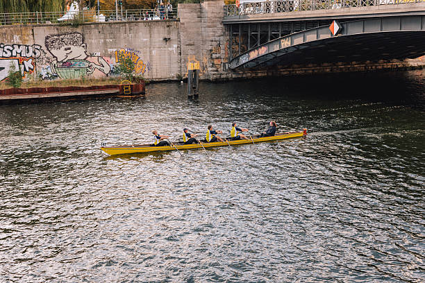 Berlin, Germany – October 11, 2025: A rowing team of five athletes moves along the Spree River in a yellow racing boat under a bridge with graffiti in central Berlin. The coxswain directs the crew as they pass through autumn reflections.