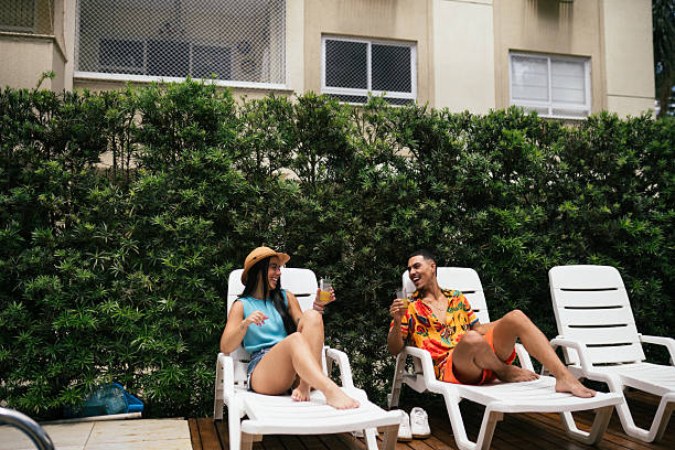 Happy young couple sitting on white lounge chairs next to a pool, enjoying juice and conversation on a sunny day.