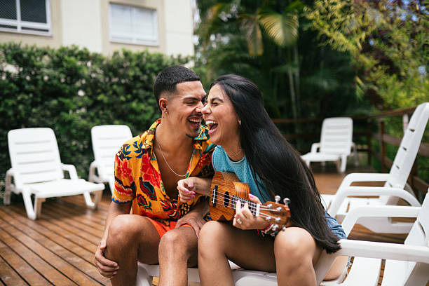 Smiling young couple sitting together on a lounge chair on a wooden deck by the pool, looking at each other while holding a small ukulele