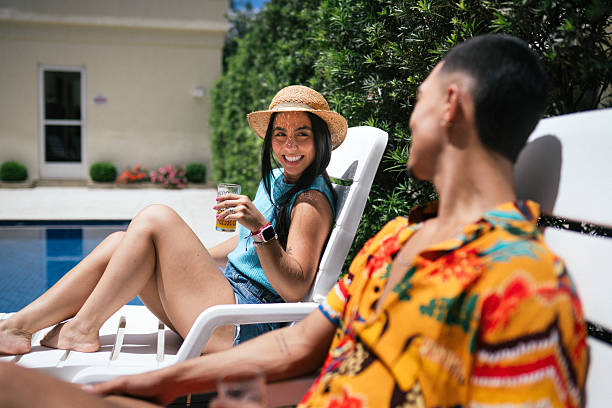 Happy young couple sitting on white lounge chairs next to a pool, enjoying juice and conversation on a sunny day.