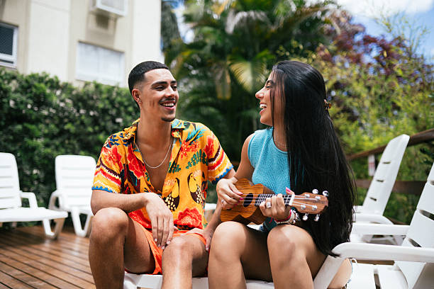 Smiling young couple sitting together on a lounge chair on a wooden deck by the pool, looking at each other while holding a small ukulele