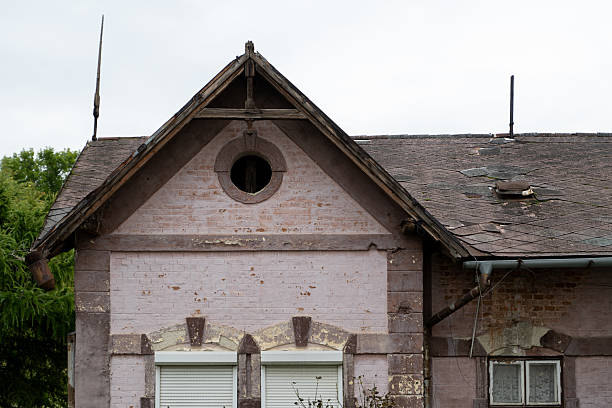 A close-up of an old brick house with a steep gable roof, featuring a circular attic window and two shuttered windows below. The weathered surface and missing roof shingles reveal the building’s age and rustic charm. The image reflects historical architecture and the quiet beauty of decay.