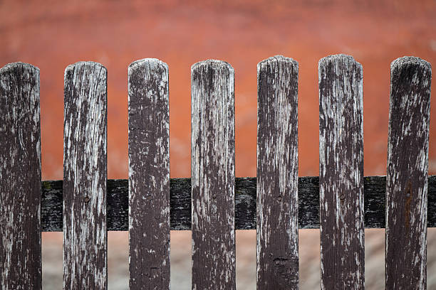 A close-up view of an old wooden fence with peeling paint and textured, weathered surfaces. Behind the dark brown planks, a softly blurred red wall creates a warm contrast. The image highlights rustic charm, aging materials, and the beauty of simple rural details.