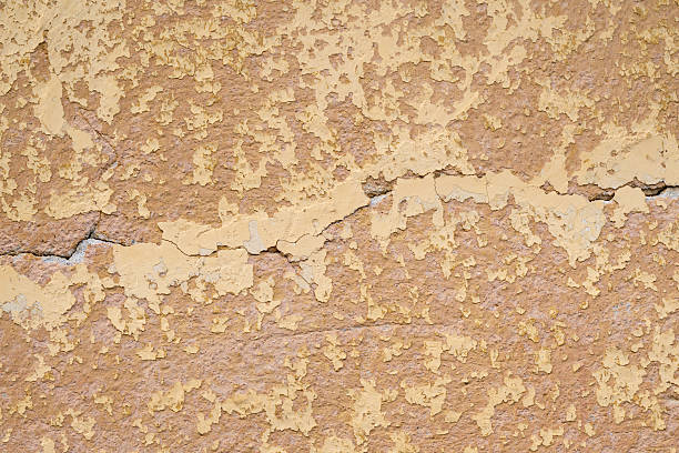 A close-up view of a yellow wall showing cracked and peeling paint on a rough plaster surface. The weathered texture and uneven color tones reveal the natural process of decay and aging. The image provides a rustic and authentic background for architectural or artistic concepts.