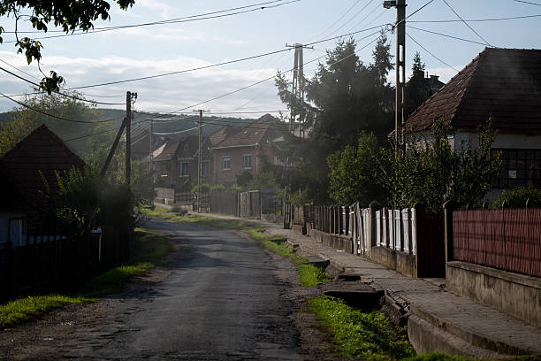 A peaceful rural street lined with old houses and garden fences illuminated by soft morning sunlight. The narrow asphalt road winds gently through the village, surrounded by greenery and calm atmosphere. The image evokes a sense of tranquility and simple countryside life.