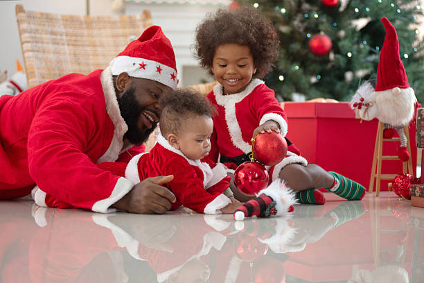 A middle-aged African father and his two sons are wearing Santa costumes playing on the floor in front of Christmas tree at home.Baby interest decorrate red ball. Christmas and New Year