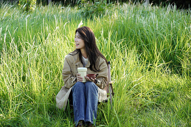 This series of images captures a woman dressed in a khaki trench coat and dark blue jeans, enjoying a serene moment in a lush field of tall green grass. Seated on a portable folding chair, she holds a thermos cup with a leather strap, gently gripping the lid while a soft smile plays on her face. Bathed in bright natural light and surrounded by vibrant greenery—with hints of foliage and faint flower buds in the background—she exudes a sense of relaxed ease. The scene perfectly embodies a slow, tranquil moment of solitude, seamlessly blending with the quiet beauty of nature.