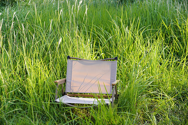 This series of images captures a woman dressed in a khaki trench coat and dark blue jeans, enjoying a serene moment in a lush field of tall green grass. Seated on a portable folding chair, she holds a thermos cup with a leather strap, gently gripping the lid while a soft smile plays on her face. Bathed in bright natural light and surrounded by vibrant greenery—with hints of foliage and faint flower buds in the background—she exudes a sense of relaxed ease. The scene perfectly embodies a slow, tranquil moment of solitude, seamlessly blending with the quiet beauty of nature.
