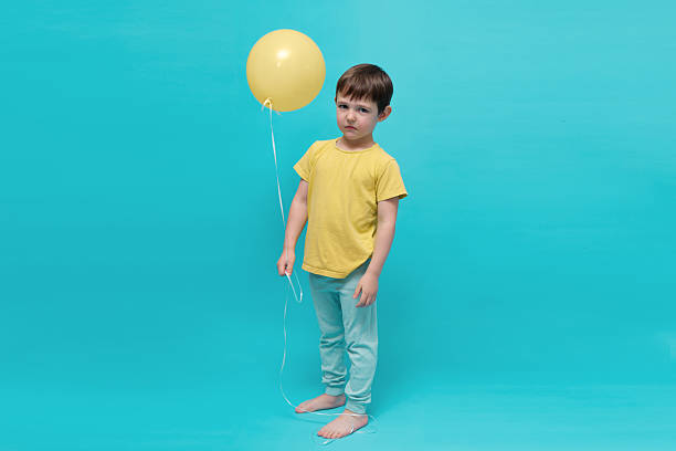 istock Little boy with a stern expression holding a yellow balloon on a blue background. Child feeling unamused or disappointed at a party. 2243529139
