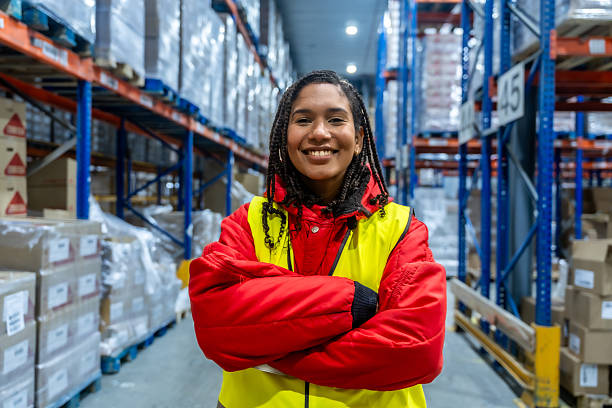 Young female employee standing confidently with crossed arms in a commercial freezer warehouse, wearing a safety vest and warm jacket, representing efficient supply chain operations
