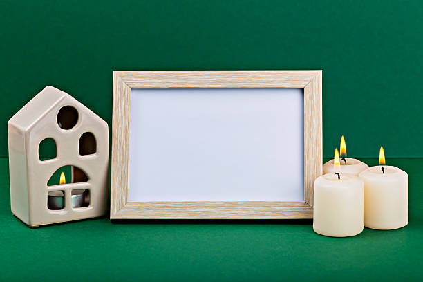 Decorative ceramic house and empty frame with lit candles on green background for cozy ambiance