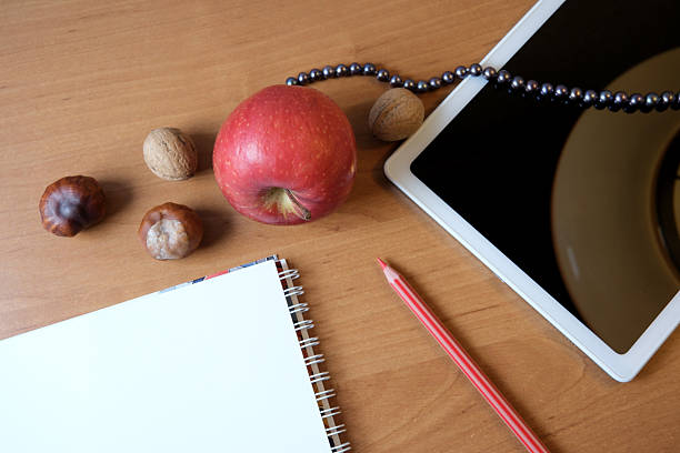 A warm wooden table setting featuring an apple, nuts, and a tablet alongside a notepad.