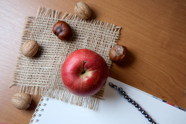 A red apple rests among walnuts on burlap, with a sketchbook nearby and a bracelet.