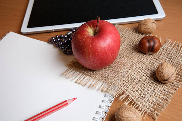 A red apple sits on a burlap cloth next to a blank sketchbook and tablet, with nuts.