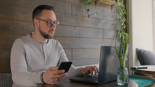 istock Man working on laptop while using smartphone in cozy cafe with plants in background 2243549828