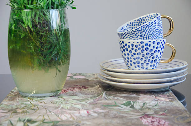 Elegant ceramic cups with golden handles and blue dotted patterns, placed on stacked plates next to a glass vase with green plants, styled on a floral surface. Perfect for kitchen decor, tableware, and lifestyle concepts.