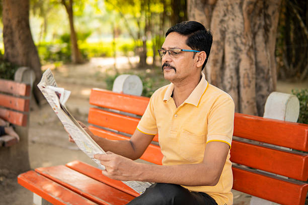 Mid adult Indian man sitting on a bench and reading a news paper in fresh air in the park at marring in leisure time.