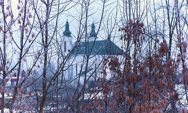 A serene winter landscape shows a church situated among leafless trees, creating a calm, snowy atmosphere in the early morning.