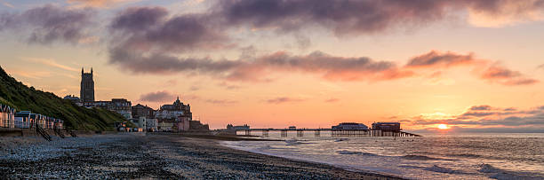 A peaceful late evening coastal landscape featuring the colourful beach huts lining the pebble and sand shoreline in Norfolk, England. Soft pastel tones fill the sky as the sun sets beyond the horizon, creating a gentle blend of oranges, pinks and gold light across the seafront. The iconic pier stretches out into the North Sea, with the historic Parish Church tower visible above the town rooftops in the distance.The steep grassy cliff and vibrant beach huts add a vivid splash of character to this traditional British seaside destination, known for its Victorian heritage, crabbing culture, and classic promenade experiences. The moody cloud formations highlight the calm yet atmospheric coastal mood often associated with the East of England shoreline.