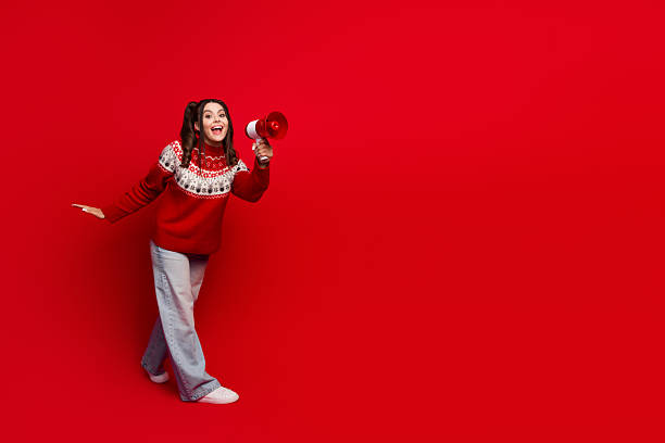 istock Young woman in a red Christmas sweater with megaphone strikes a dynamic pose against a solid red background promoting holiday fun 2243571846