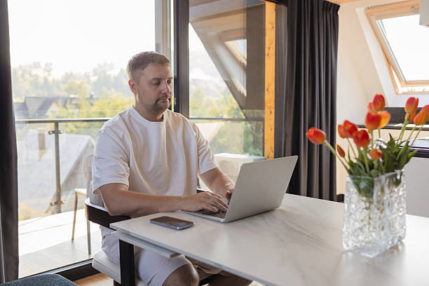 istock Man working on laptop at home office with flowers on table. . Modern workspace with natural light, tulips on the table and cozy atmosphere. 2243572126