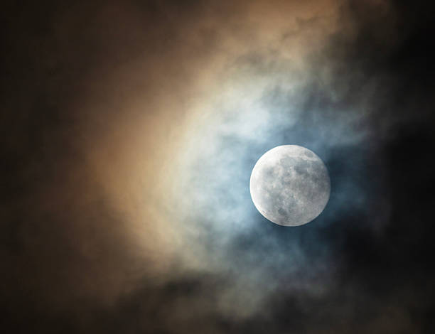 A thin patch of clouds on an overcast night is letting the full moon shine through and casting orange and blue colors above.