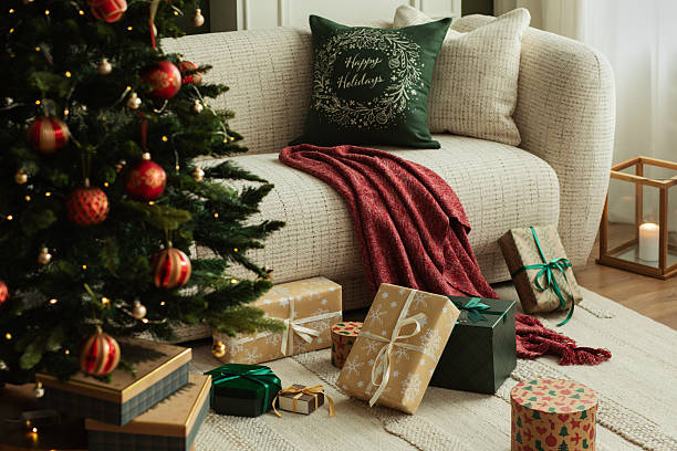 Christmas living room with decorated tree and presents under it. Cozy beige sofa, red throw blanket and stylish holiday decorations.