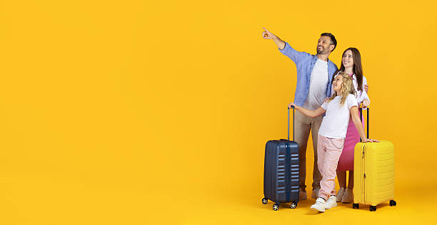 istock Smiling family standing with suitcases on yellow background, pointing at copy space 2243587318