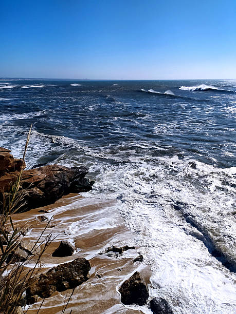 istock Dramatic coastal scene showcasing powerful ocean waves crashing against a sandy shore with rocky formations under a clear blue sky. Perfect for travel, nature, and resilience themes. 2243587724