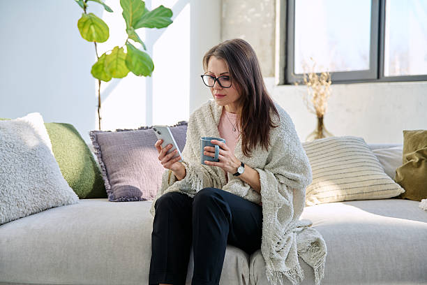 istock Woman under blanket with mug of hot drink using mobile phone sitting on sofa 2243597287