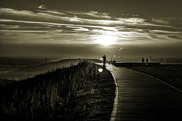 Where the sky kisses the earth, there is a path through the silence. Immersed in Sepia, this scene lies there like an old memory - the cyclist a lonely wanderer between day and night, the silhouettes on the horizon silent witnesses of a fleeting moment. The sun breaks through layers of clouds like liquid gold, strips of light on the way and makes the world stop. It is these intermediate moments - not day, not night, not arrival, not departure - in which we feel that the path itself is the goal.